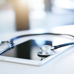 Kuvituskuva, jossa on valkoinen tablet-tietokone pöydällä ja sen päällä musta stetoskooppi.
