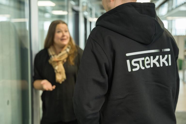Kuvituskuva, jossa yhdellä henkilöllä on musta Istekki-huppari päällään ja hän seisoo selin kameraan päin. Hän juttelee naishenkilön kanssa toimiston käytävällä.