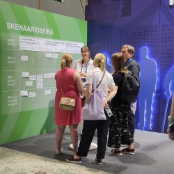 Istekin ATK-päivien ständi ja sen skenaarioseinä. Skenaarioseinä on väriltään omenanvihreä. Skenaarioseinän edessä on joukko ihmisiä keskustelemassa Istekin edustajan kanssa tulevaisuuden skenaarioista.